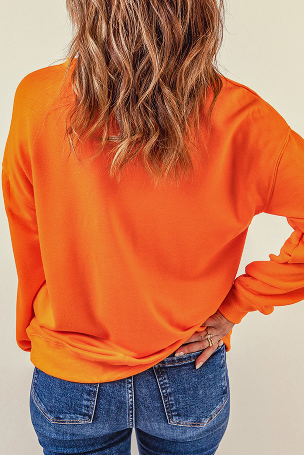 Person with wavy, shoulder-length hair wearing an Orange Plain Crew Neck Pullover Sweatshirt and blue jeans, standing with hand on hip, facing away.