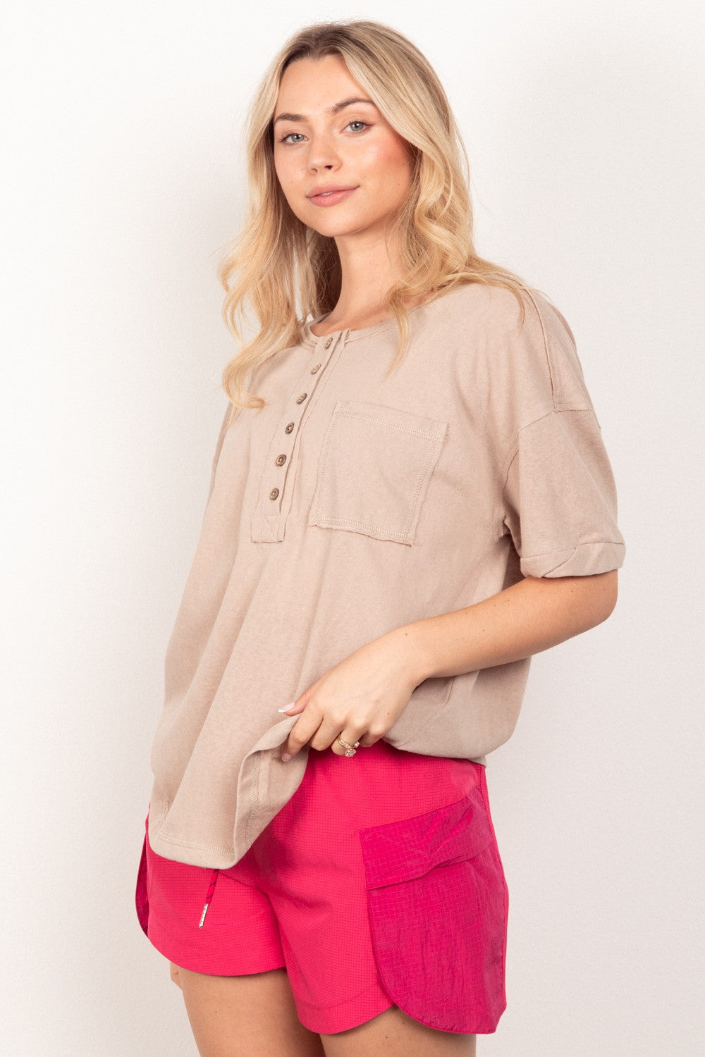 A woman with long blonde hair wears the VERY J Twisted Sleeve Band Half Button Top and pink shorts, reminiscent of a 90s-inspired wardrobe, standing against a plain white background.