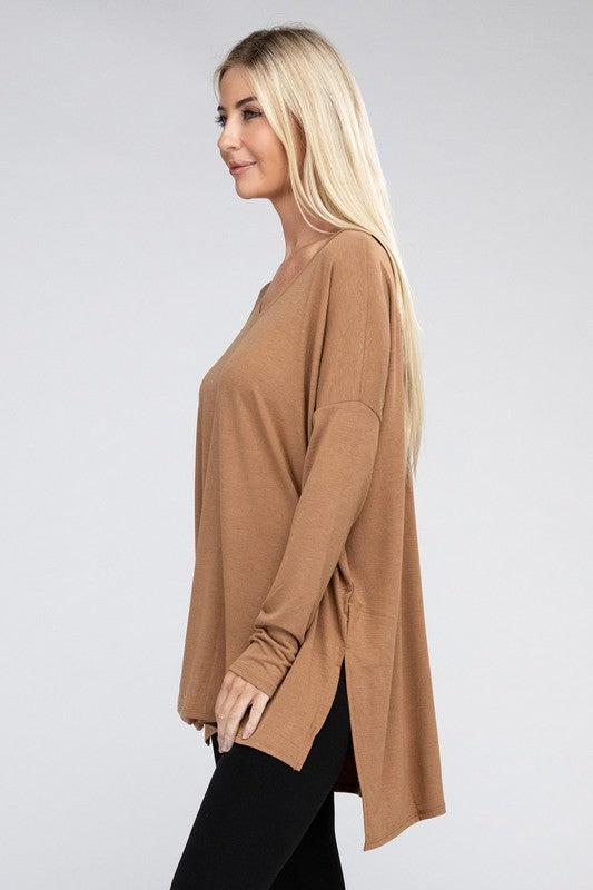 A woman with long, blonde hair is wearing a loose-fitting, brown Dolman Long Sleeve V-Neck Side Slit Hi-Low Hem Top and black pants. She poses with one hand on her hip and a neutral facial expression against a light grey background.