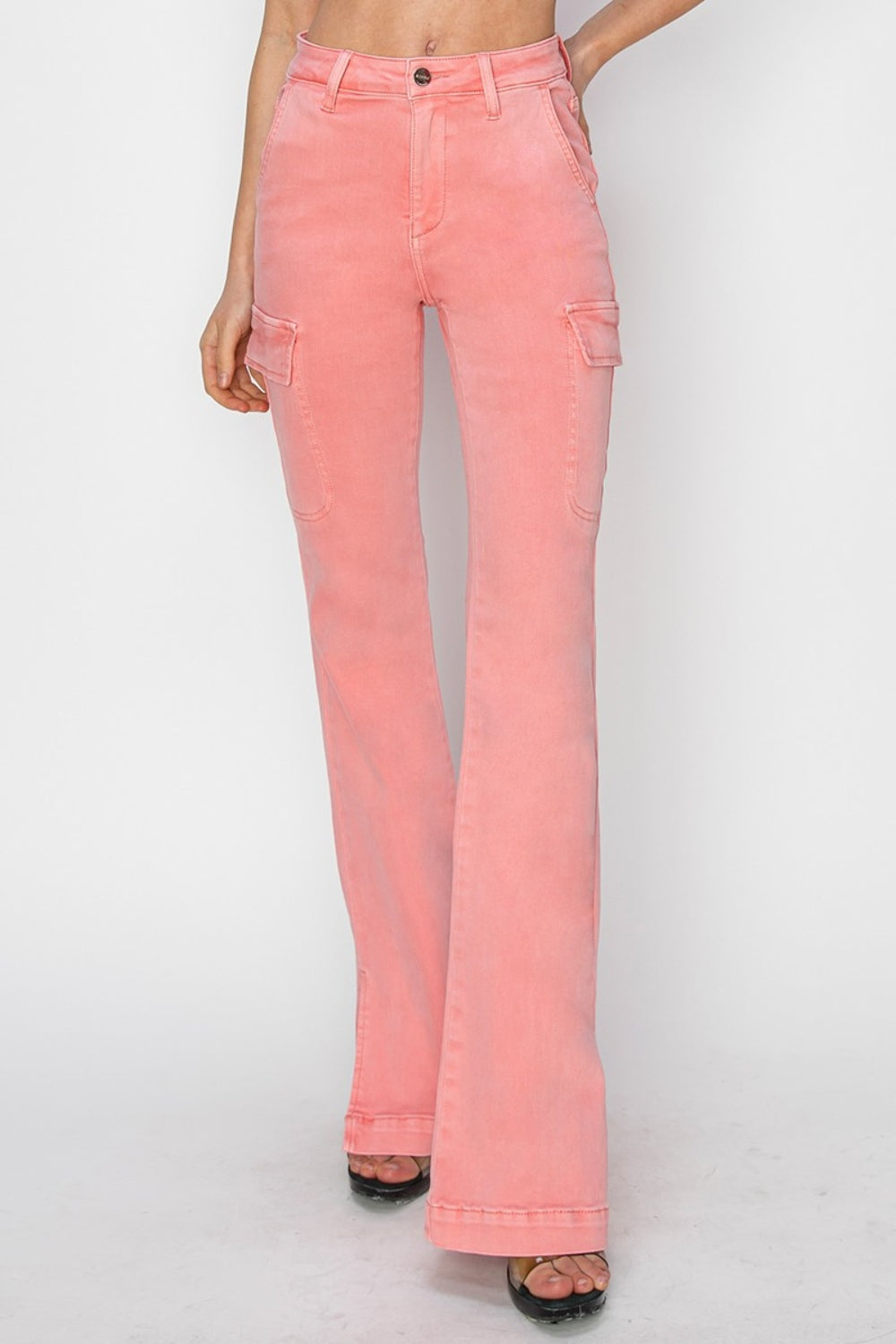 A person wearing RISEN Full Size High Rise Side Slit Cargo Bootcut Jeans, featuring a pink flared design with front pockets, stands against a plain background.