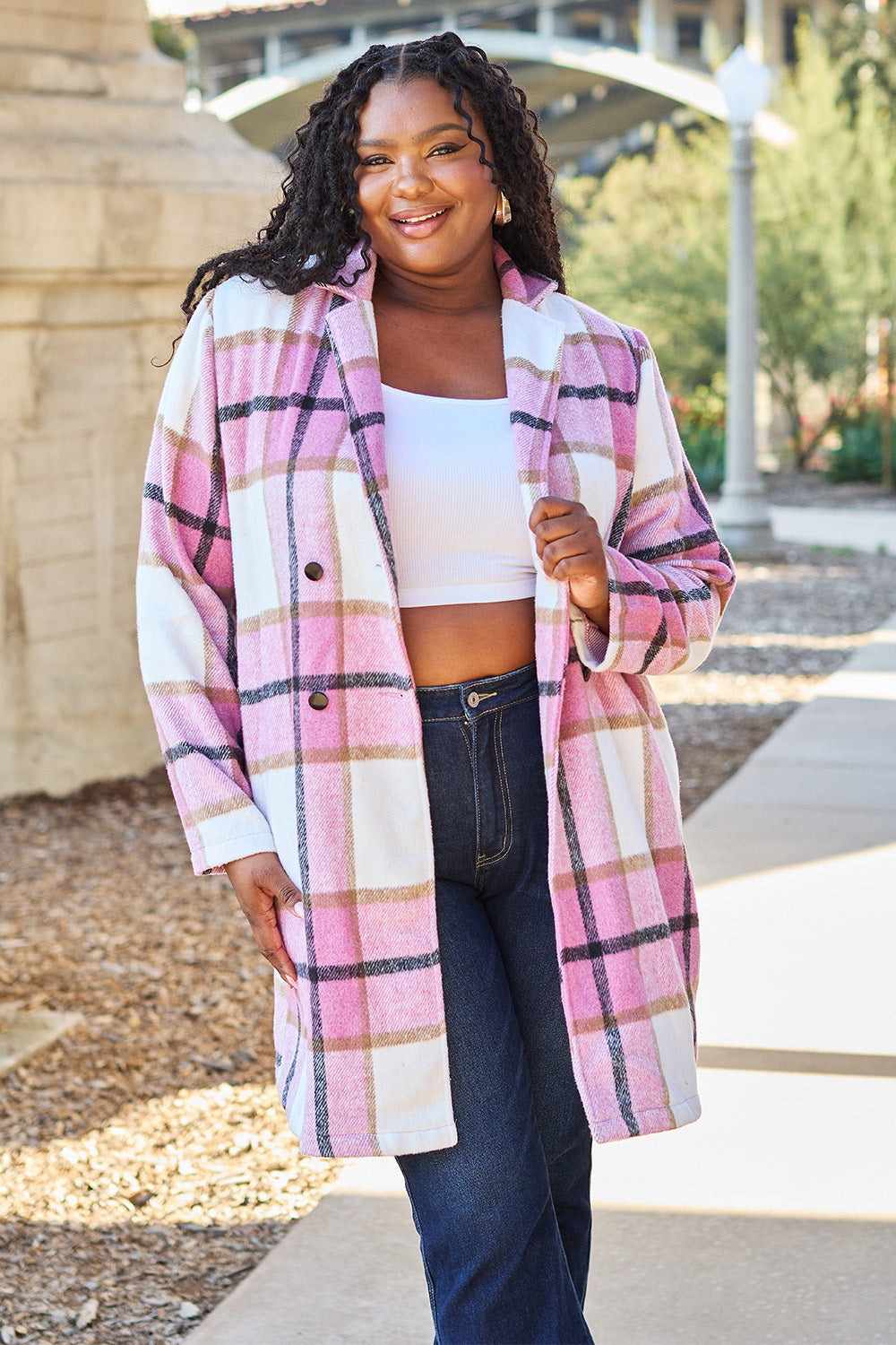 A woman stands on a sidewalk wearing the Double Take Full Size Plaid Button Up Lapel Collar Coat in pink and white over a white top and blue jeans, with her right hand lightly touching her hair. The polyester fabric of the coat makes it durable and perfect for an easy machine wash cold.