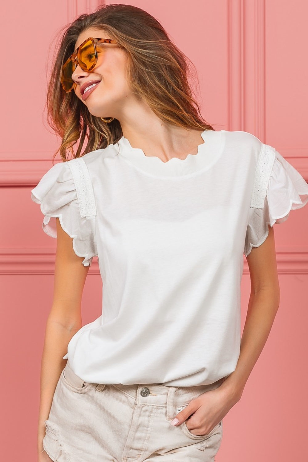 A woman with wavy hair dons the elegant BiBi Scallop Edged Lace Trim Top and large amber sunglasses while standing in front of a vibrant pink wall.