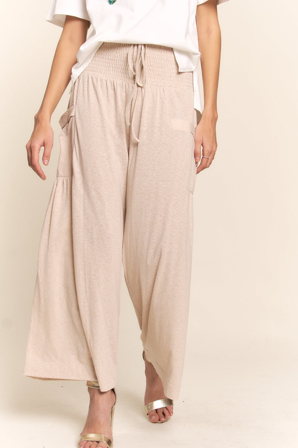 A person wearing J.NNA Smocked Waist Boho Wide Leg Pants with Pockets in beige stands in low-heeled sandals against a plain background, paired with a chic white T-shirt.