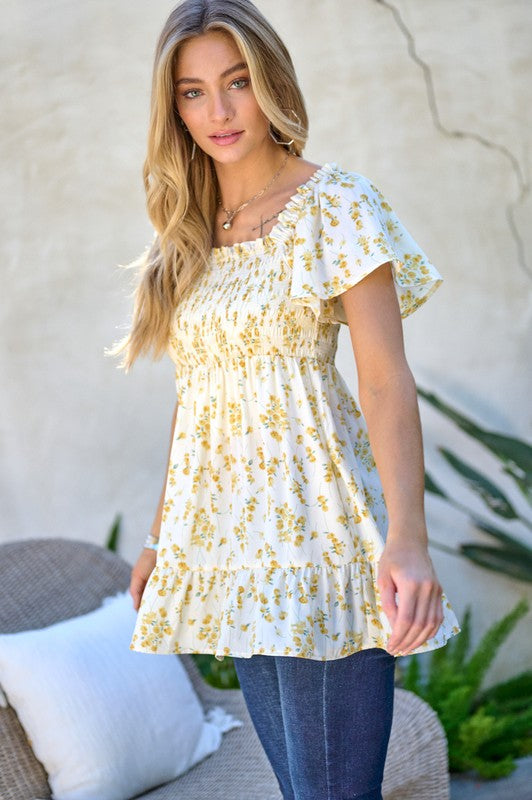 A woman stands outdoors donning a Floral Printed V-Neck Ruffle Top paired with jeans, set against a neutral background, with plants in the foreground, highlighting her fashion-forward wardrobe.