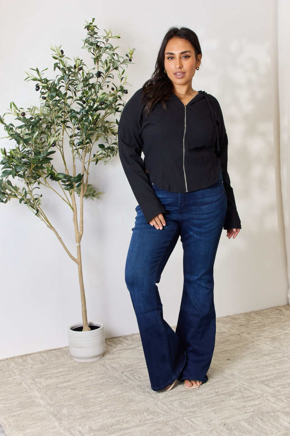 A person wearing the Culture Code Full Size Ribbed Zip Up Drawstring Hooded Jacket and blue jeans stands in front of a plain background with a small leafy plant to the left.