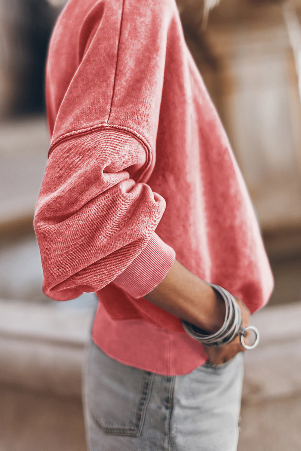 A person is wearing a Drop Shoulder Crew Neck Pullover Sweatshirt in a coral color with sleeves rolled up and bracelets on their wrist, paired with light blue jeans. The drop shoulder design adds a relaxed vibe to the look. The background is blurred.