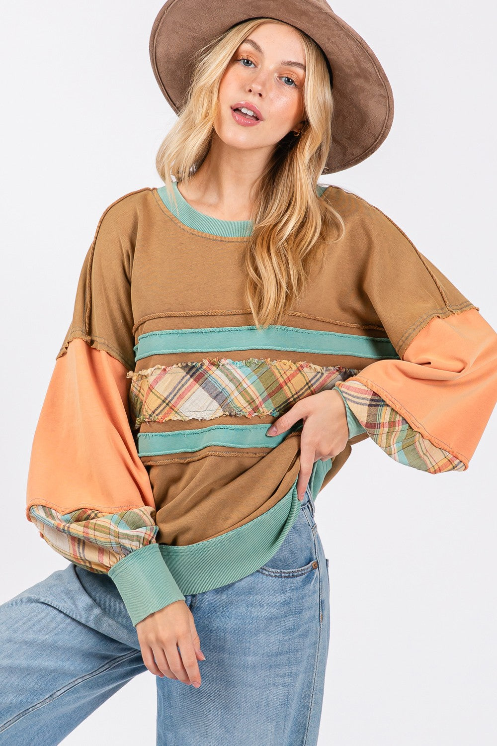 A person wearing a brown hat and the SAGE + FIG Mineral Wash Raw Edge Color Block Sweatshirt, featuring a patchwork design with plaid print contrast and solid color panels, smiles while posing with one hand in their pocket.