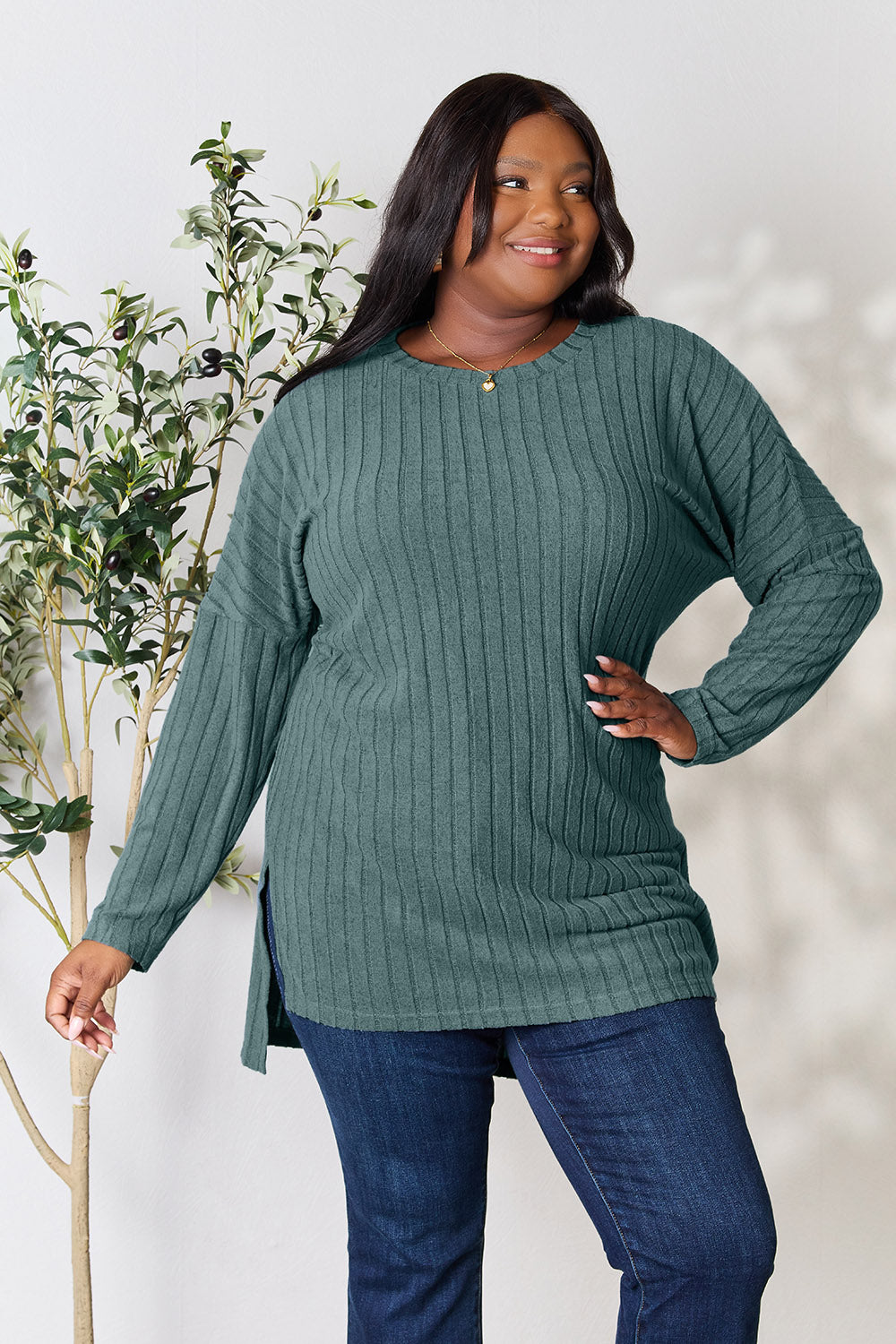 A person with shoulder-length hair smiles while wearing a beige ribbed sweater called the Basic Bae Full Size Ribbed Round Neck Long Sleeve Slit Top, along with matching beige pants, showcasing a basic style. They stand next to a green plant.