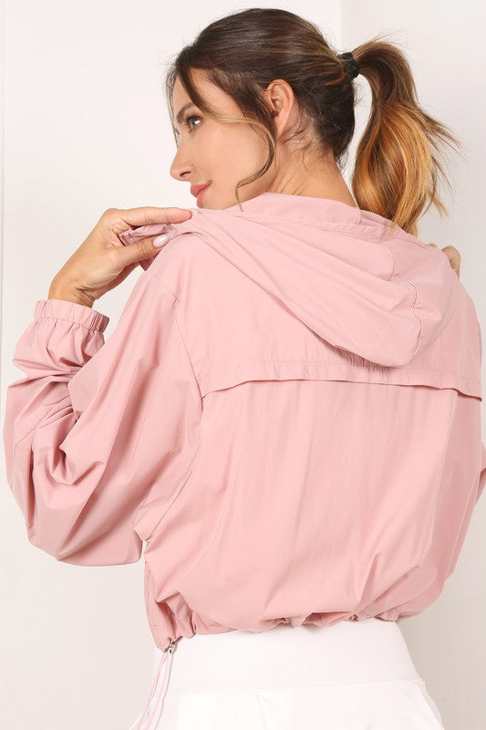 A woman wearing a pink wind breaker over a white top stands against a white wall.