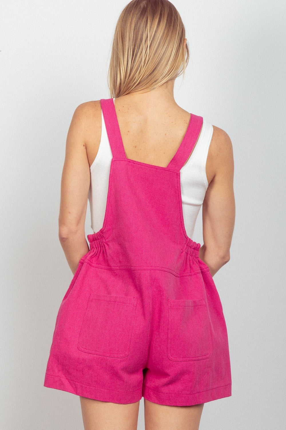 A woman stands against a plain background, smiling, wearing stylish VERY J Adjustable Suspender Overalls with Pockets in pink overalls layered over a white tank top.