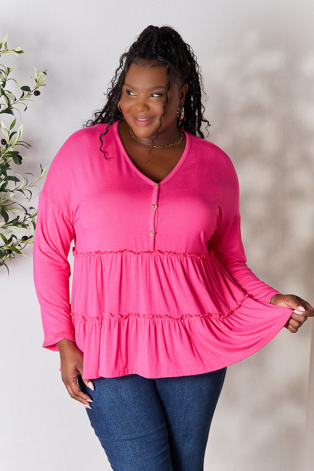 Person stands indoors near a plant, wearing the Double Take Half Button Long Sleeve Ruffle Hem Blouse in gray and blue jeans, smiling while looking to the side. The slightly stretchy fabric of the blouse adds comfort while its buttoned design offers a touch of elegance.