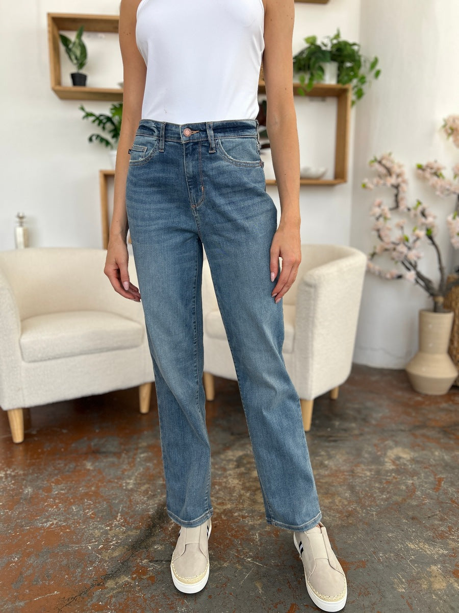 Person wearing Judy Blue Full Size High Waist Straight Jeans, a white shirt, and black sandals stands on a polished concrete floor with a white armchair in the background.