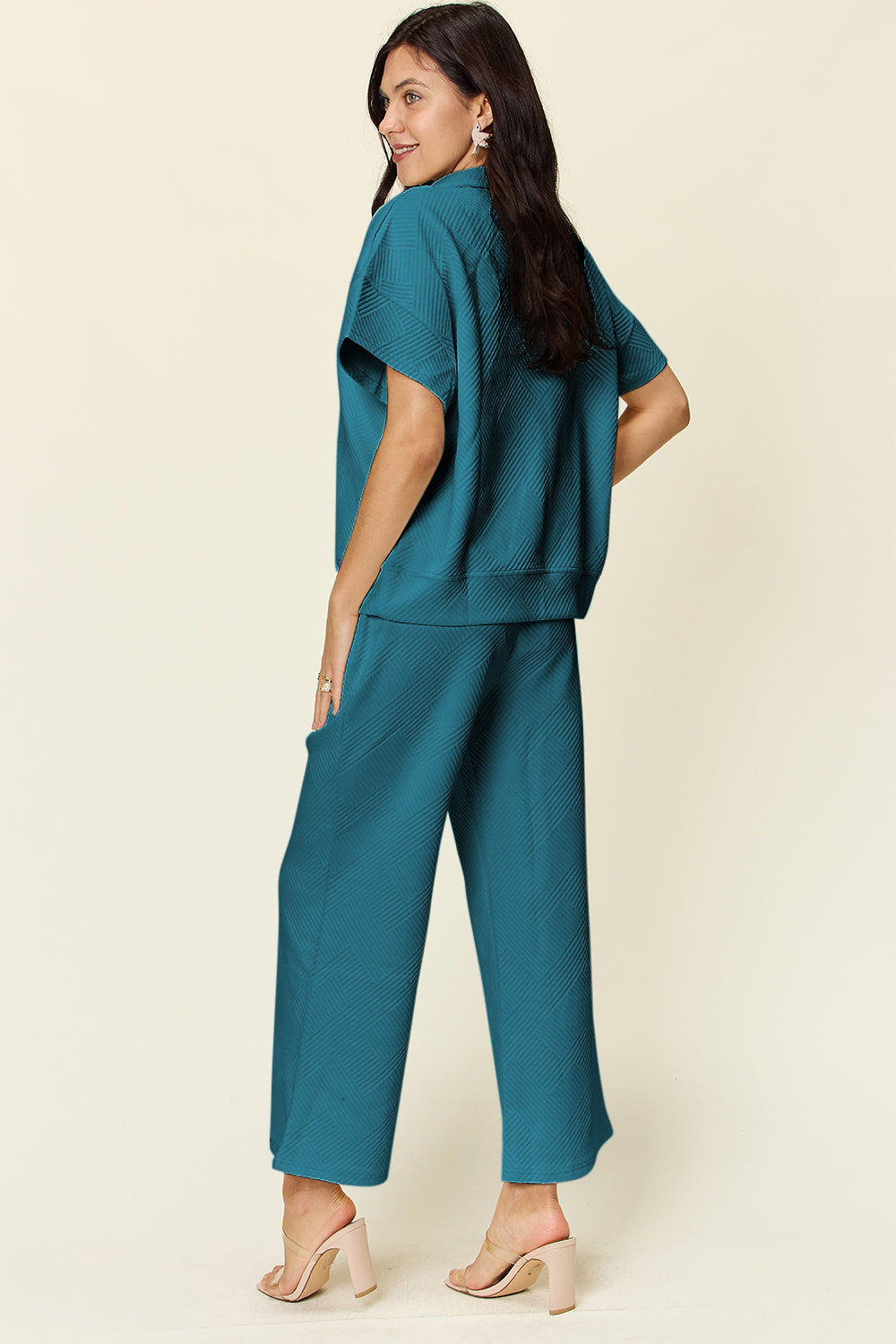 A woman is wearing the Double Take Full Size Texture Half Zip Short Sleeve Top and Pants Set, featuring a white zip-up short-sleeve top and matching wide-leg, drawstring pants. She stands with one hand in her pocket and smiles against a light background.