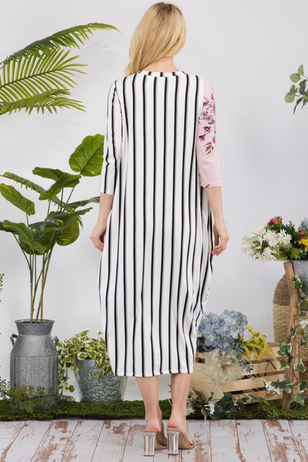 A woman stands amidst potted plants and flowers on a wooden floor, wearing the elegant and chic Celeste Full Size Floral Striped Contrast Midi-Dress with Pockets.