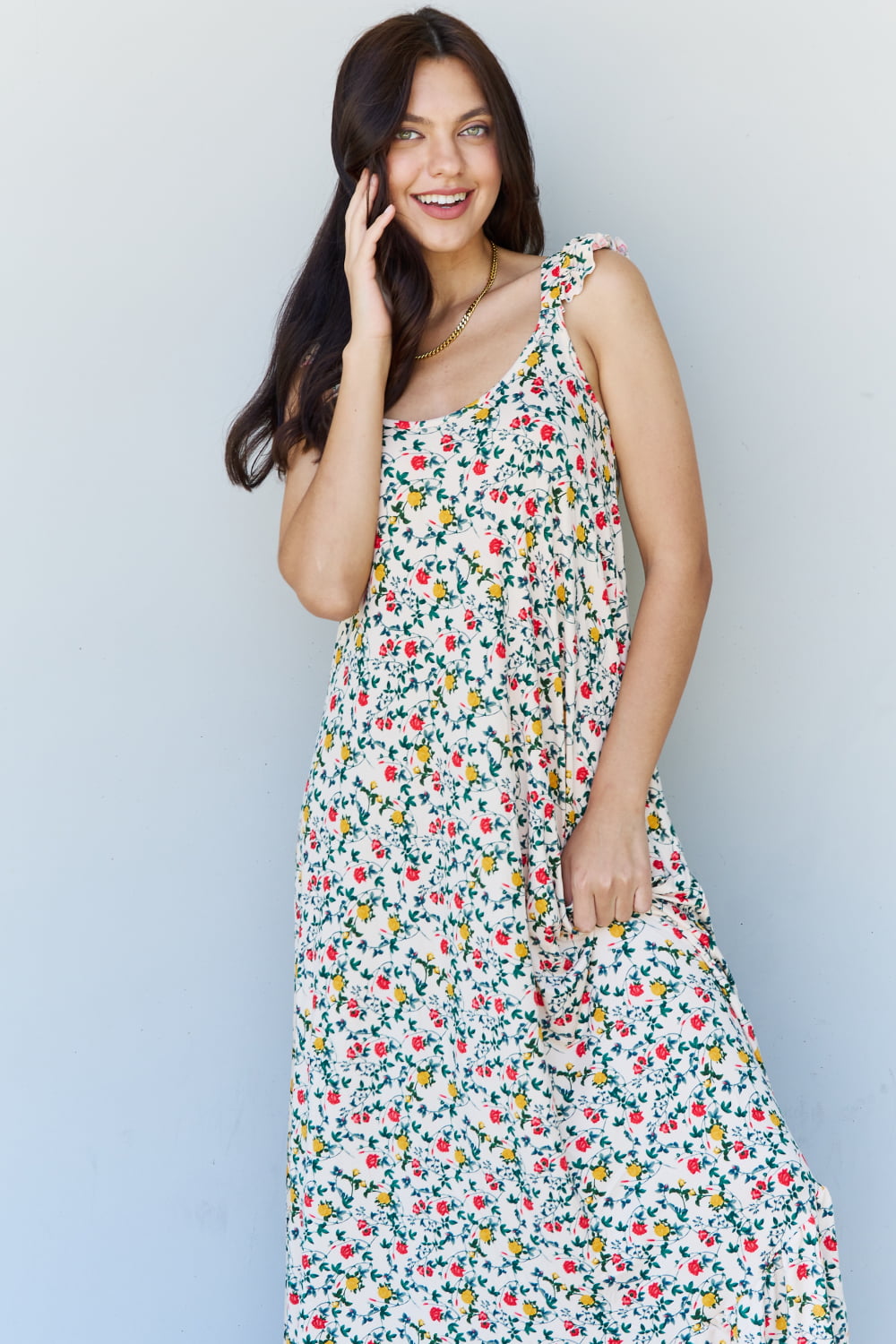 A woman is standing against a gray wall, wearing the Doublju In The Garden Ruffle Floral Maxi Dress in Natural Rose and black sandals, with her left hand raised to her head.