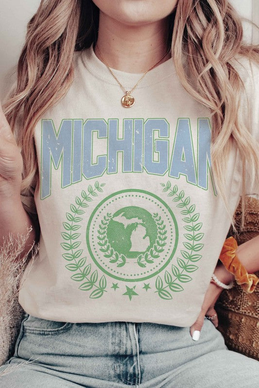 An individual dons a white MICHIGAN Graphic Tee, crafted from 100% cotton. The shirt prominently displays the word "Michigan" in blue, alongside an Earth graphic encircled by laurel leaves and topped with a star. Its unisex sizing guarantees a comfortable fit for all wearers.