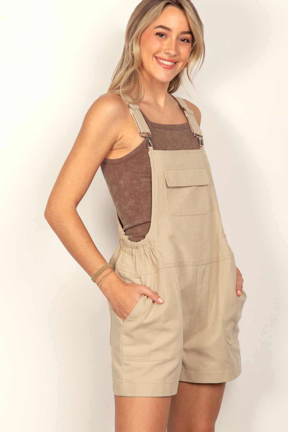 A woman wearing the VERY J Adjustable Suspender Overalls with Pockets in beige and a brown tank top smiles while touching her hair with one hand and has her other hand in a pocket.