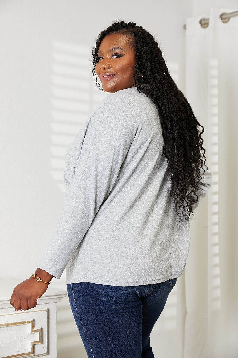 A person with long hair wearing a Double Take Seam Detail Round Neck Long Sleeve Top in solid black and blue jeans stands with one hand in their pocket, looking downward and smiling.