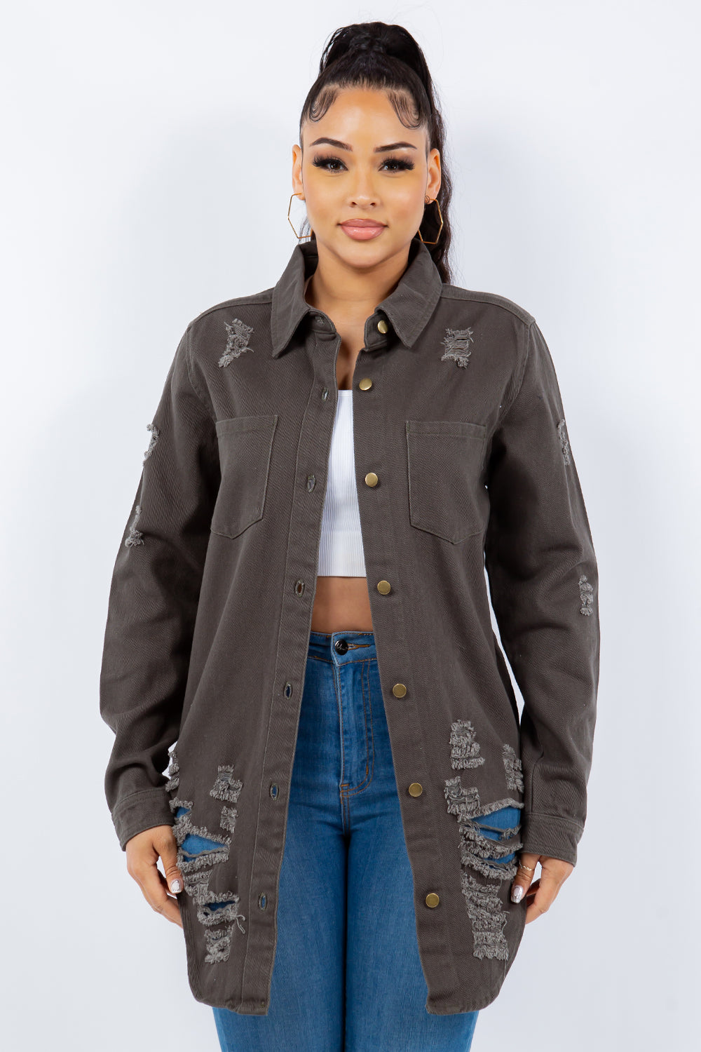 A woman stands against a light background wearing the American Bazi Distressed Button Up Long Sleeve Denim Jacket with a white crop top and blue ripped jeans.