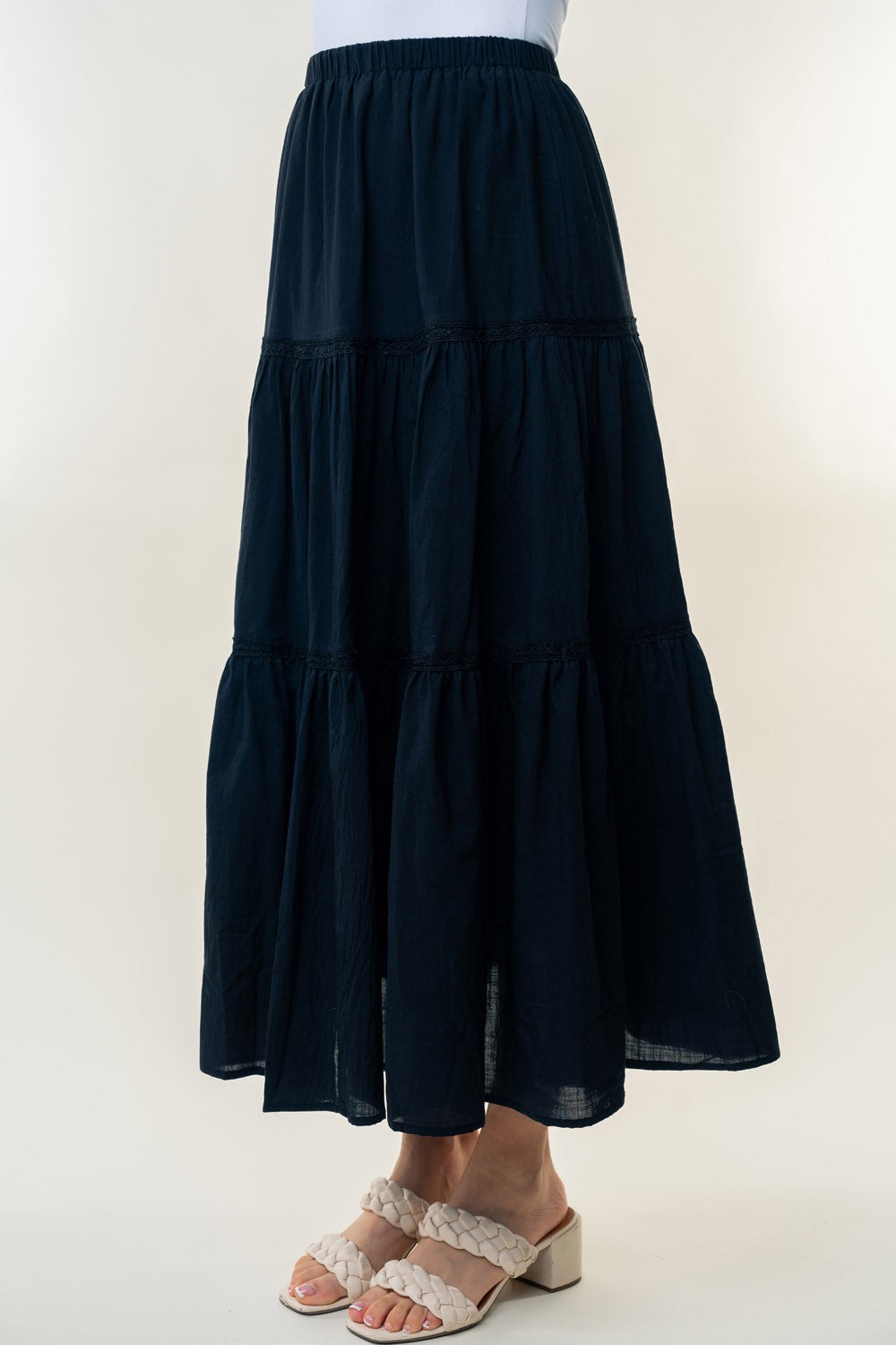 A long, White Birch High Waisted Tiered Maxi Skirt in navy blue, offering a versatile piece perfect for a bohemian look. It's worn by a person paired with white heeled sandals. The background is plain and light-colored.