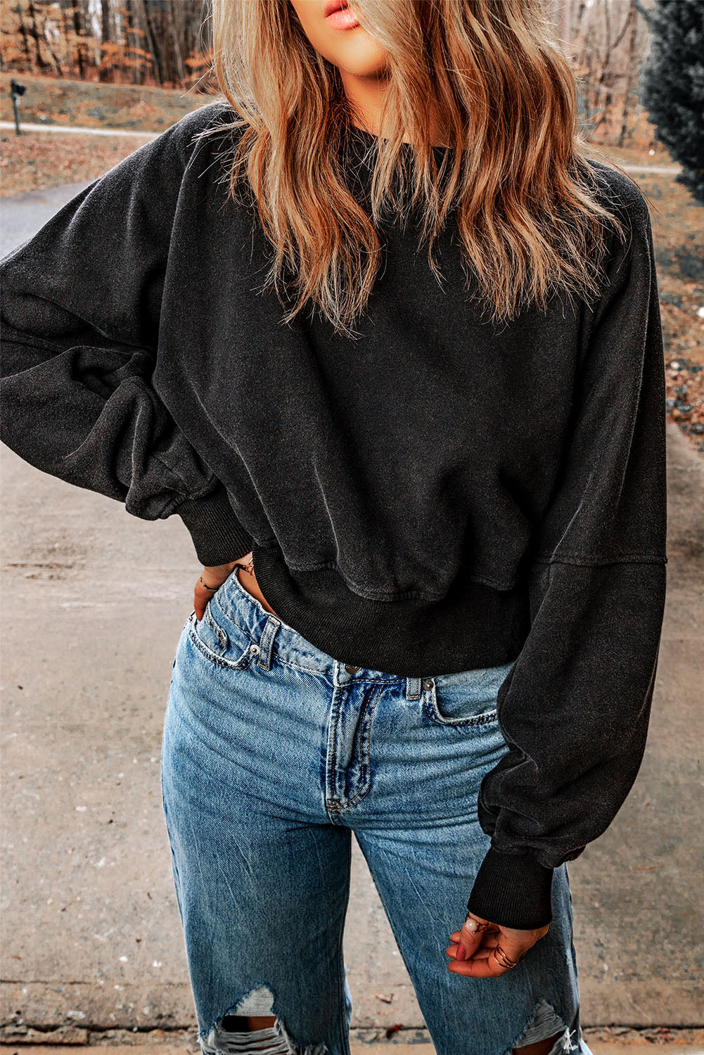 A person with wavy, shoulder-length hair wearing a Black Acid Wash V-shape Open Back Sweatshirt and distressed blue jeans stands outside on a paved surface.