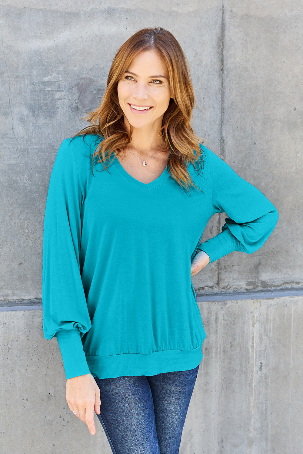 A woman with brown hair is standing against a concrete wall, wearing the Basic Bae Full Size V-Neck Lantern Sleeve Top in teal and blue basic-style jeans, smiling.