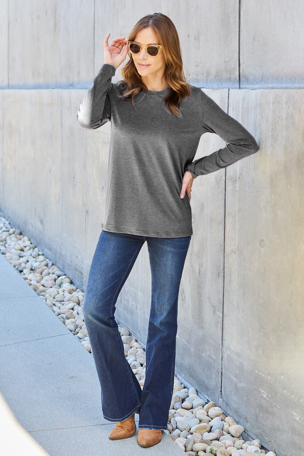 A woman with shoulder-length hair wearing the Basic Bae Full Size Round Neck Long Sleeve Top and blue jeans stands in front of a concrete wall, holding a beige handbag over her shoulder. Her outfit exemplifies basic style, with the top made from 100% polyester and recommended for machine wash cold.