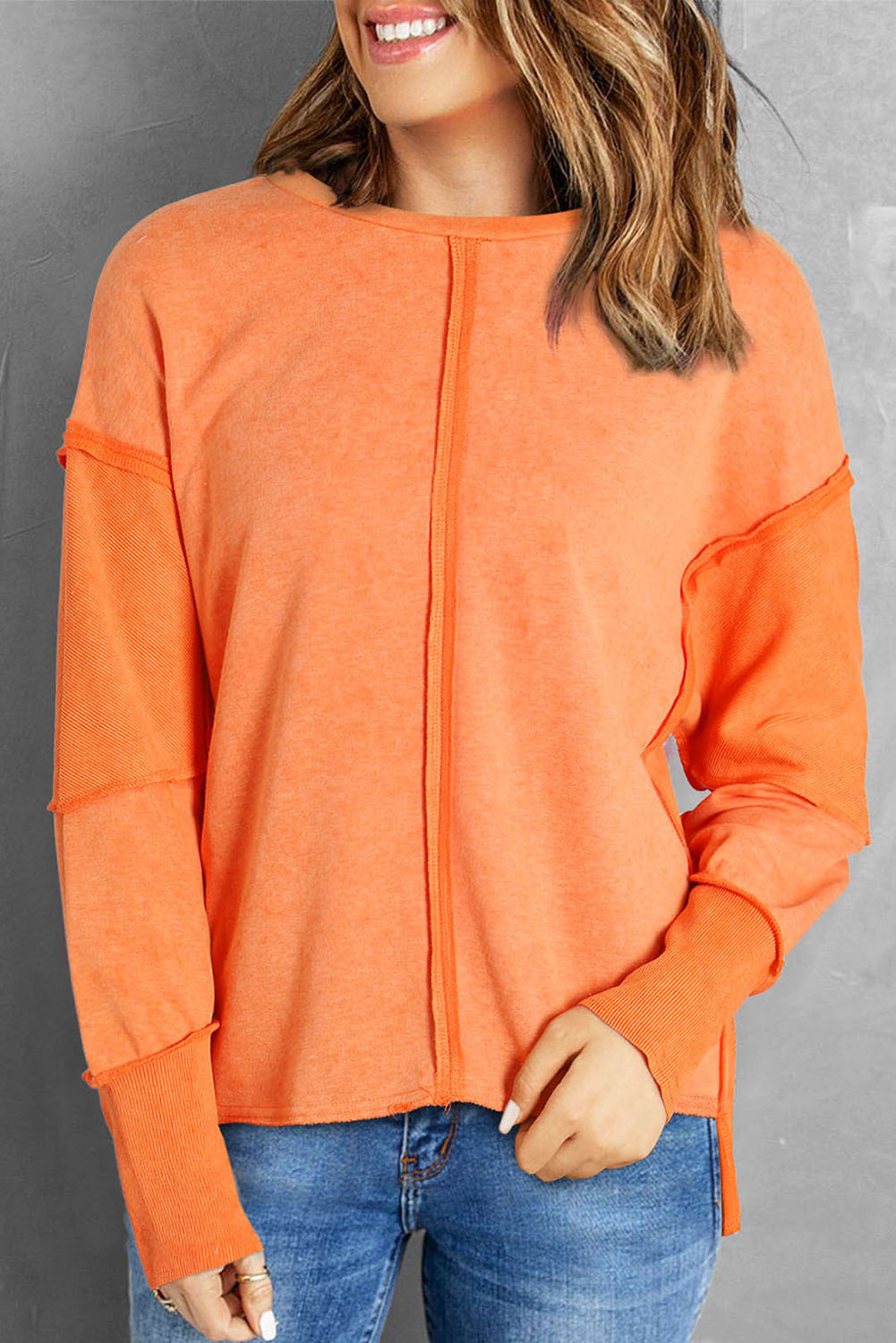 A person with long blonde hair is standing with their back to the camera, wearing an Orange Exposed Seamed High Low Raw Edge Sweatshirt and denim shorts, in front of a brick wall.