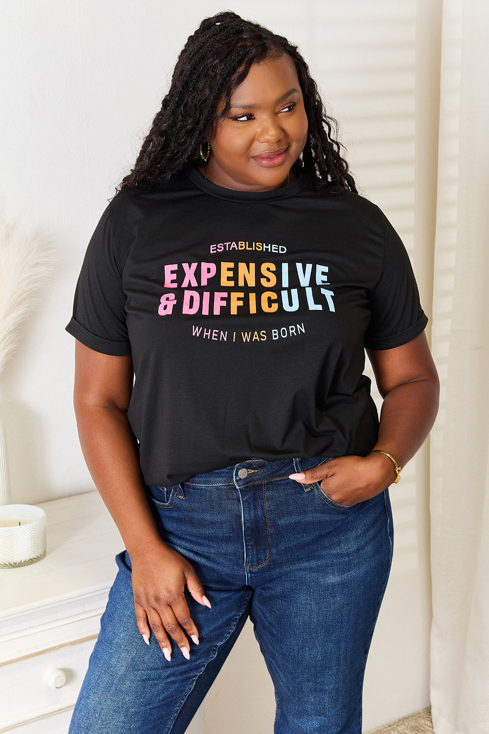 A woman with long braided hair, wearing the Simply Love Slogan Graphic Cuffed Sleeve T-Shirt that says "Established Expensive & Difficult When I Was Born," stands with one hand in her pocket. The trendy cuffed sleeves of her shirt add an extra touch of style.