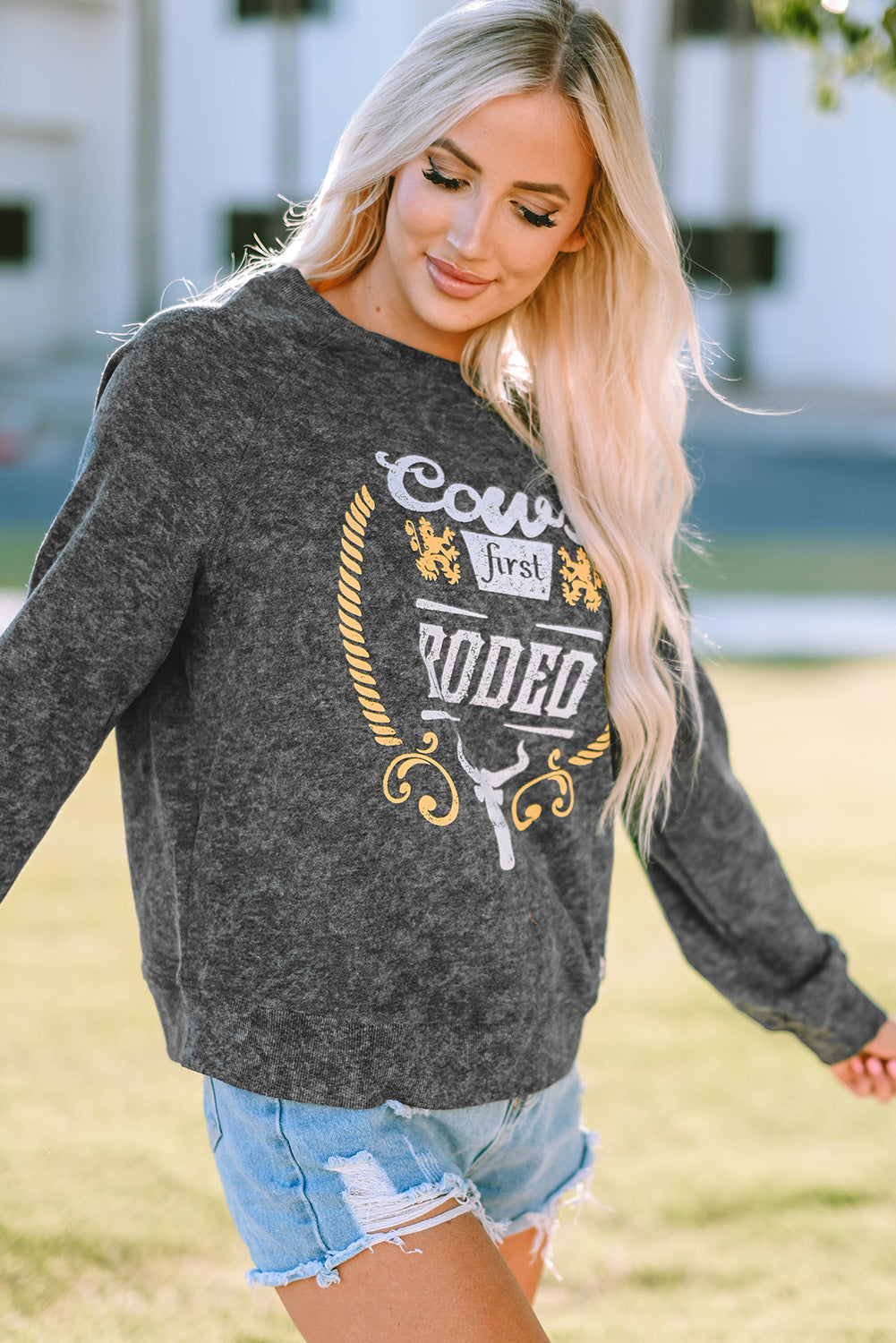 A blonde woman wearing a Gray Coors Banquet RODEO Graphic Mineral Washed Sweatshirt and light blue denim shorts, showcasing casual outdoor wear, stands outdoors with her back to the camera.