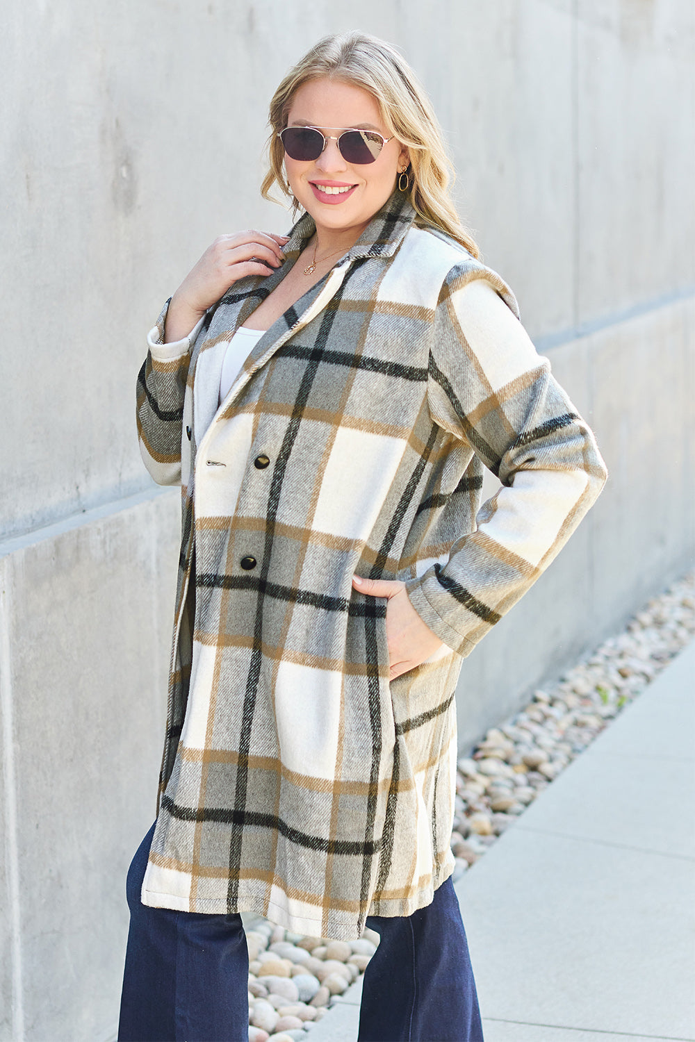 A woman stands on a sidewalk wearing the Double Take Full Size Plaid Button Up Lapel Collar Coat in pink and white over a white top and blue jeans, with her right hand lightly touching her hair. The polyester fabric of the coat makes it durable and perfect for an easy machine wash cold.