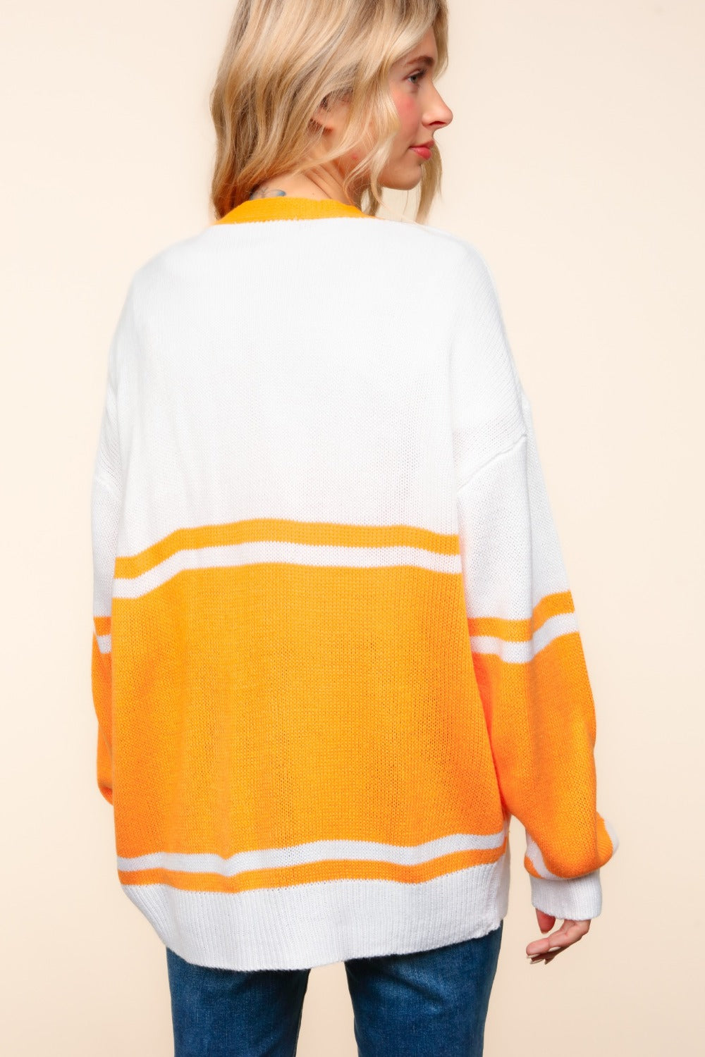 A person wearing the Haptics V Neck Button Down Letter Patch Cardigan, oversized with white and orange stripes and a yellow "T" patch, paired with blue jeans, smiles while lifting their hands to their head.