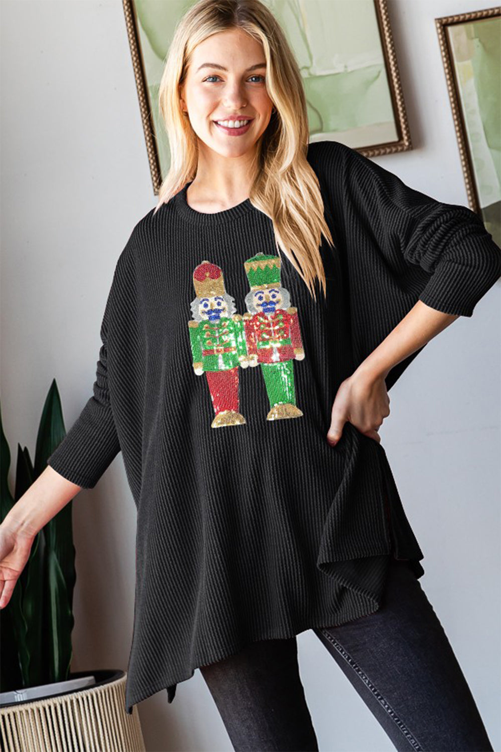 A woman wearing a black Heimish Sequin Nutcracker Long Sleeve Ribbed Top adorned with colorful figures on the front is standing indoors.