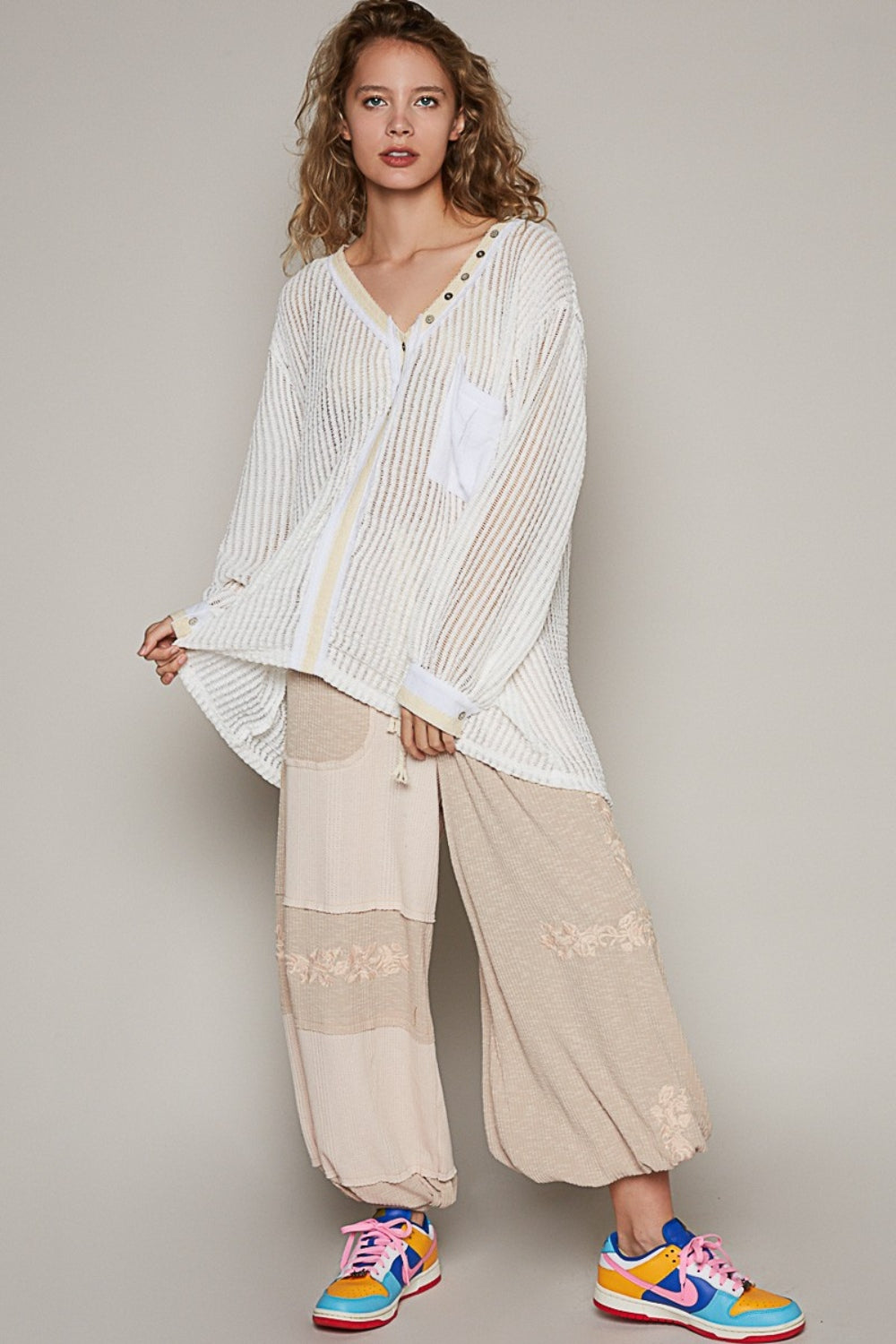 A person with wavy hair wearing a loose, bohemian-style POL V-Neck Long Sleeve Crochet Top in a sheer, striped white design paired with beige pants stands against a plain background.