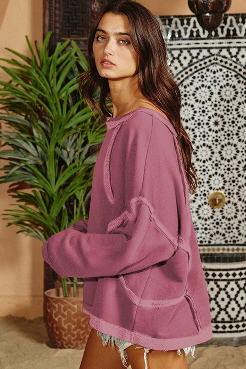 A woman with long hair is wearing an Exposed Seam Drop Shoulder Raw Hem Oversized Sweatshirt in pink, paired with denim shorts, standing indoors in front of a potted plant and a decorative wall.