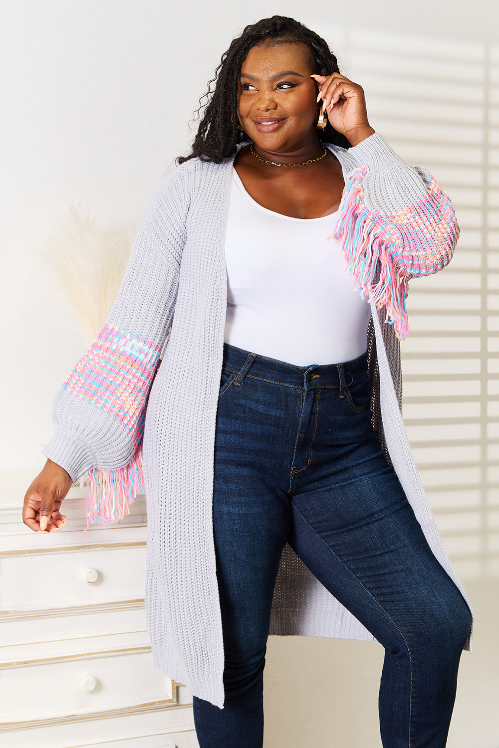 A woman poses indoors wearing the Woven Right Fringe Sleeve Dropped Shoulder Cardigan, a cozy, light gray piece featuring colorful tasselled sleeves. She pairs it with a white top and dark jeans.