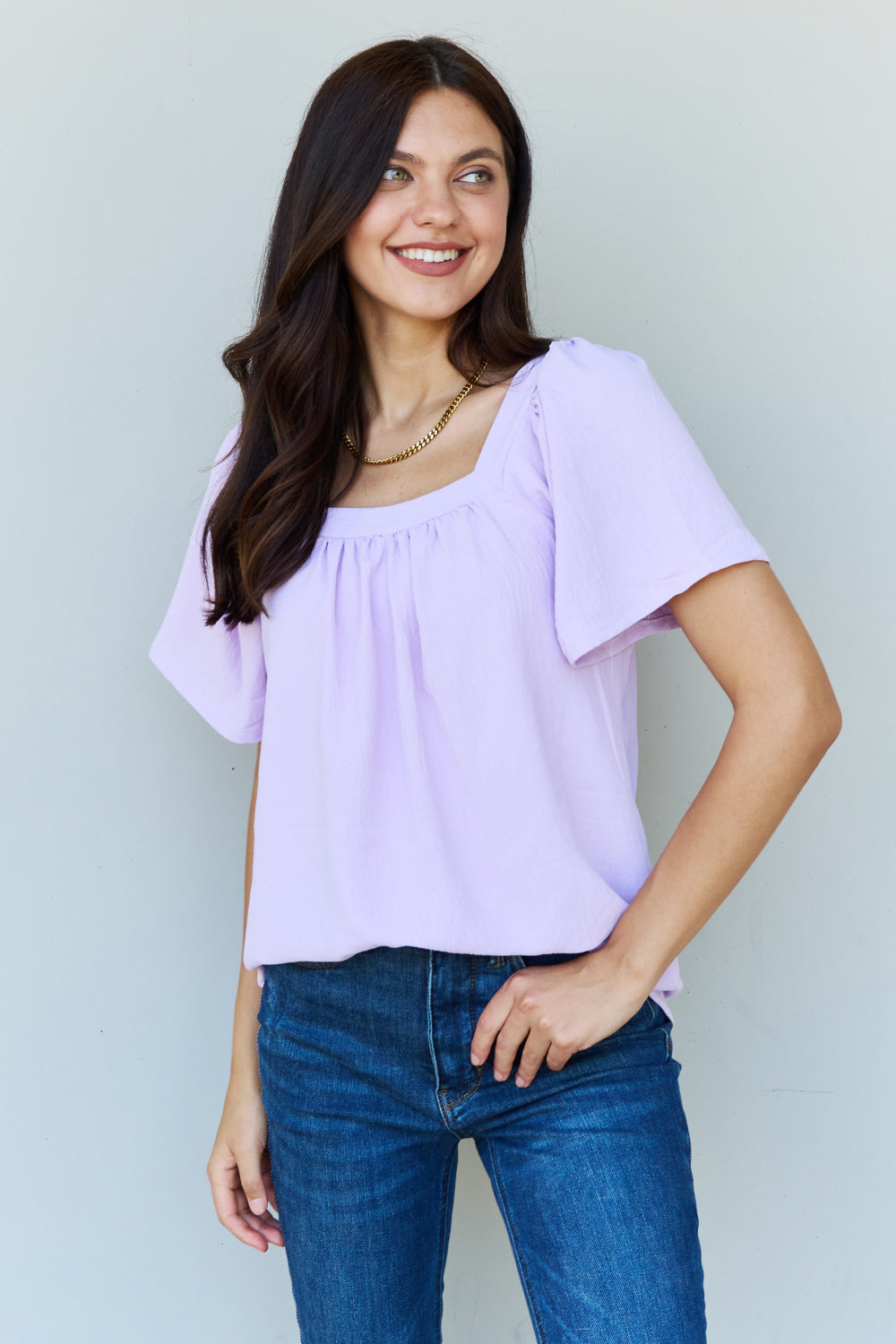 A person stands smiling in a lavender Ninexis Keep Me Close Square Neck Short Sleeve Blouse, paired with blue jeans against a plain background.