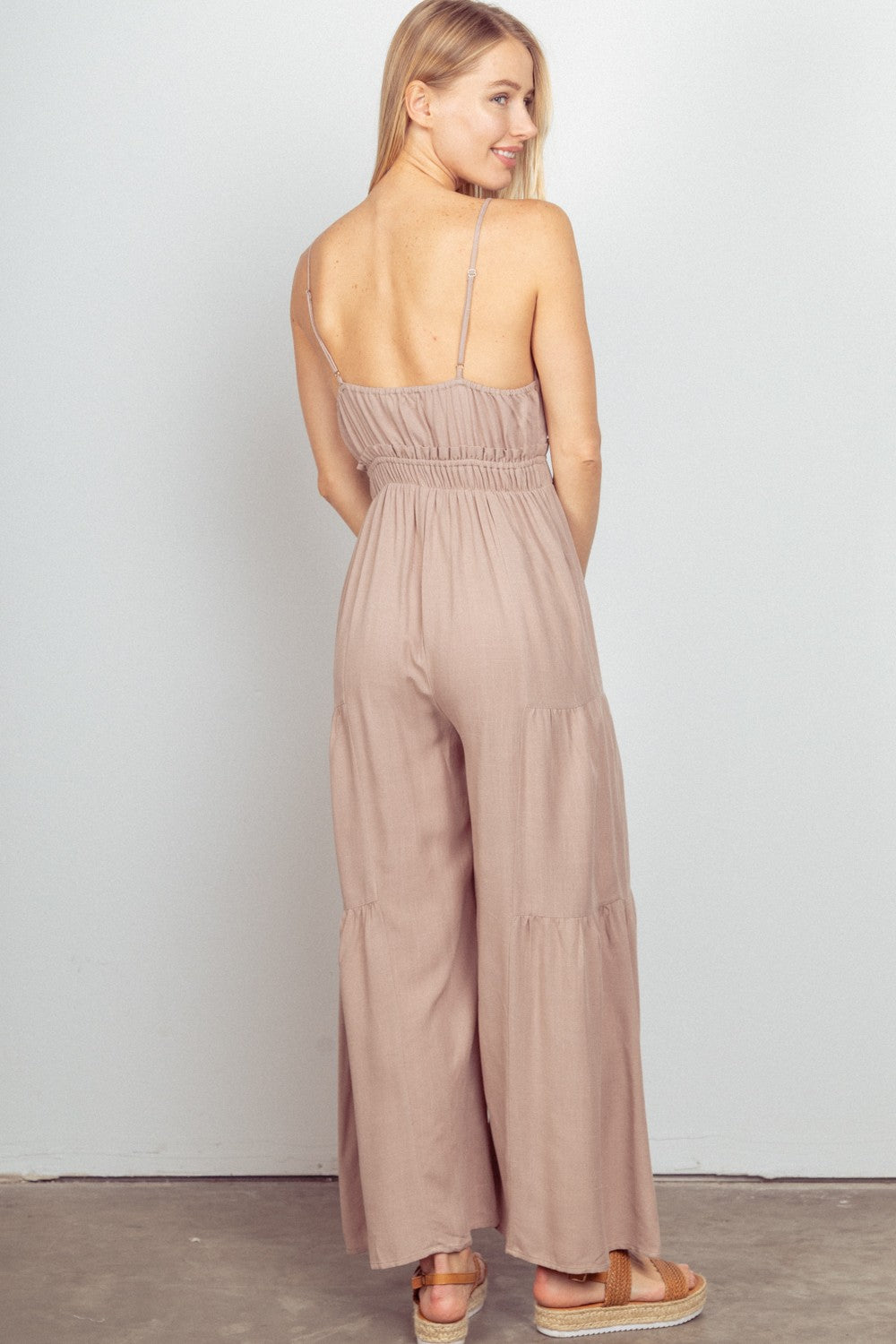 A woman standing indoors wearing the VERY J Sleeveless Ruched Wide Leg Jumpsuit in beige with adjustable straps and sandals, set against a light gray background.