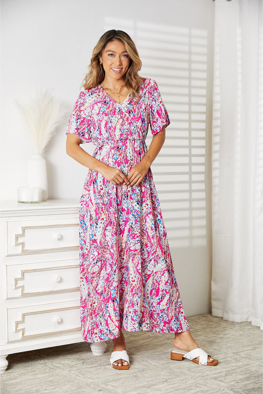 A woman stands in a room wearing the Double Take Multicolored V-Neck Maxi Dress, paired with white sandals. A dresser and curtain create a charming backdrop.