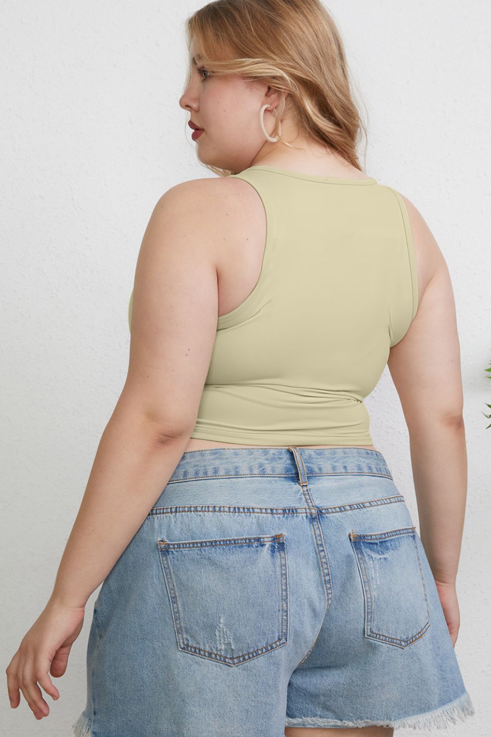 A woman with light hair wears the Basic Bae Full Size Round Neck Slim Tank in black and blue jeans, standing against a light-colored curtain.