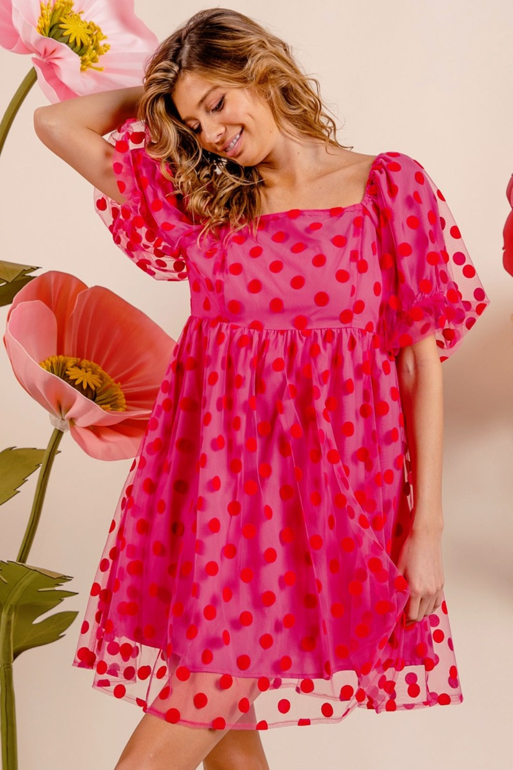 Wearing the BiBi Polka Dot Mesh Puff Sleeve Dress, a woman poses gracefully with one hand on her head. The vintage-inspired pink dress adorned with red polka dots enhances her elegance as she stands amidst large artificial flowers.