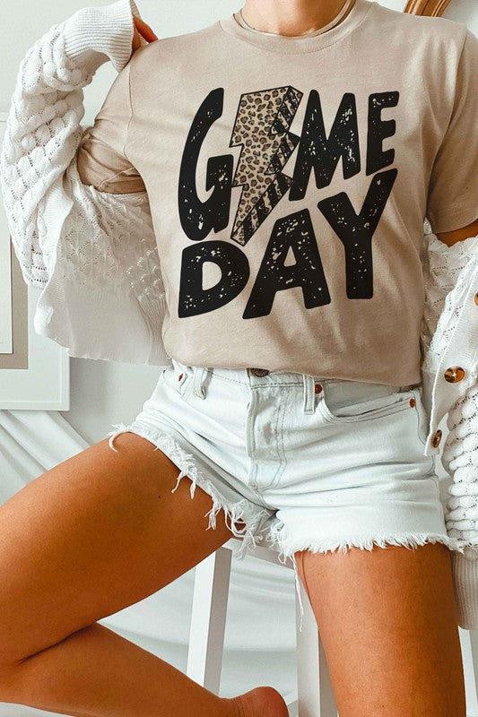 A person wearing a white "LEOPARD LIGHTNING GAME DAY Graphic Tee" with a leopard print lightning bolt design stands with a denim jacket partially on, showcasing its unisex sizing.