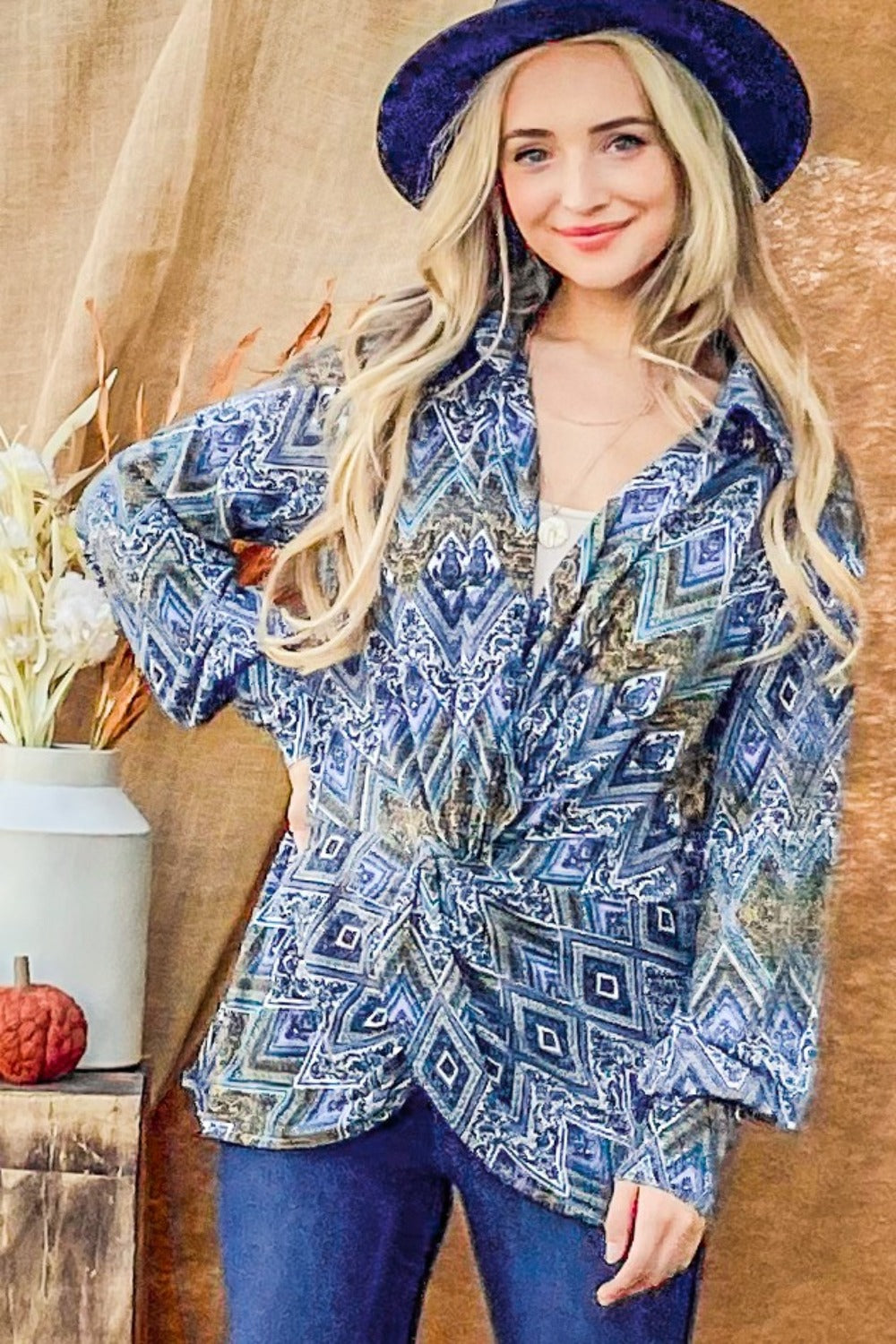 A woman with long blonde hair wearing an And The Why Print Twist Knot Long Sleeve Blouse, blue jeans, and a dark blue hat stands in front of a brown backdrop.