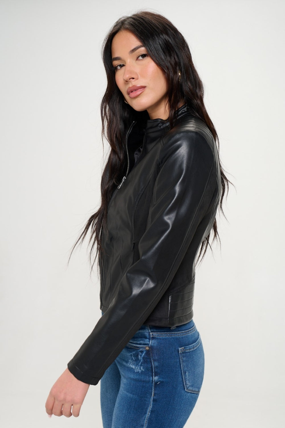 A person with long hair wearing a black Coalition LA Zip Up Vegan Moto Jacket and blue jeans stands against a plain background, showcasing a stylish spring wardrobe.