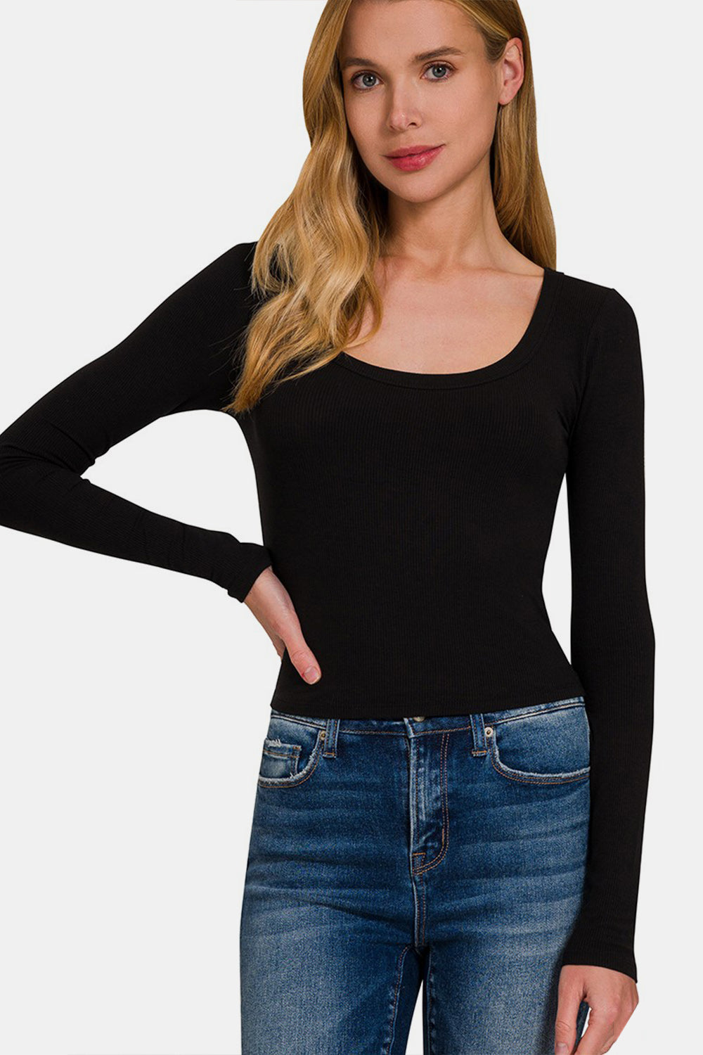 A woman with long blonde hair, wearing a black Zenana Scoop Neck Long Sleeve T-Shirt and blue jeans, stands with one hand on her hip, looking at the camera.