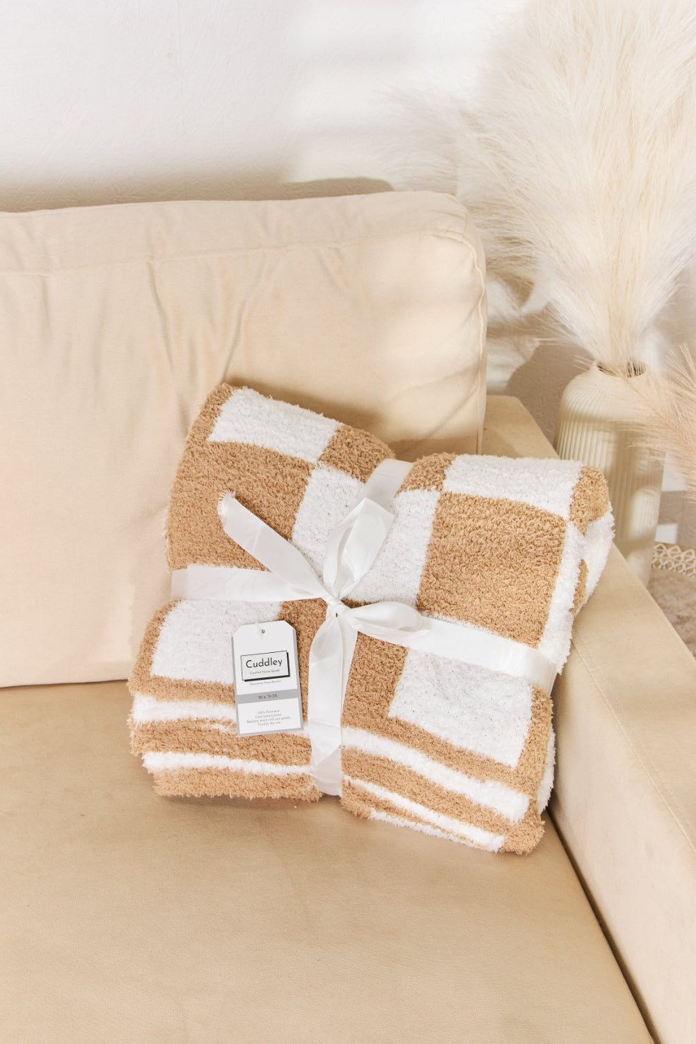 A person stands holding a large Cuddley Checkered Decorative Throw Blanket, smiling, in a room with a sofa and a potted plant. The blanket features a black and white checkerboard pattern, is made from 100% polyester, and imported for quality.