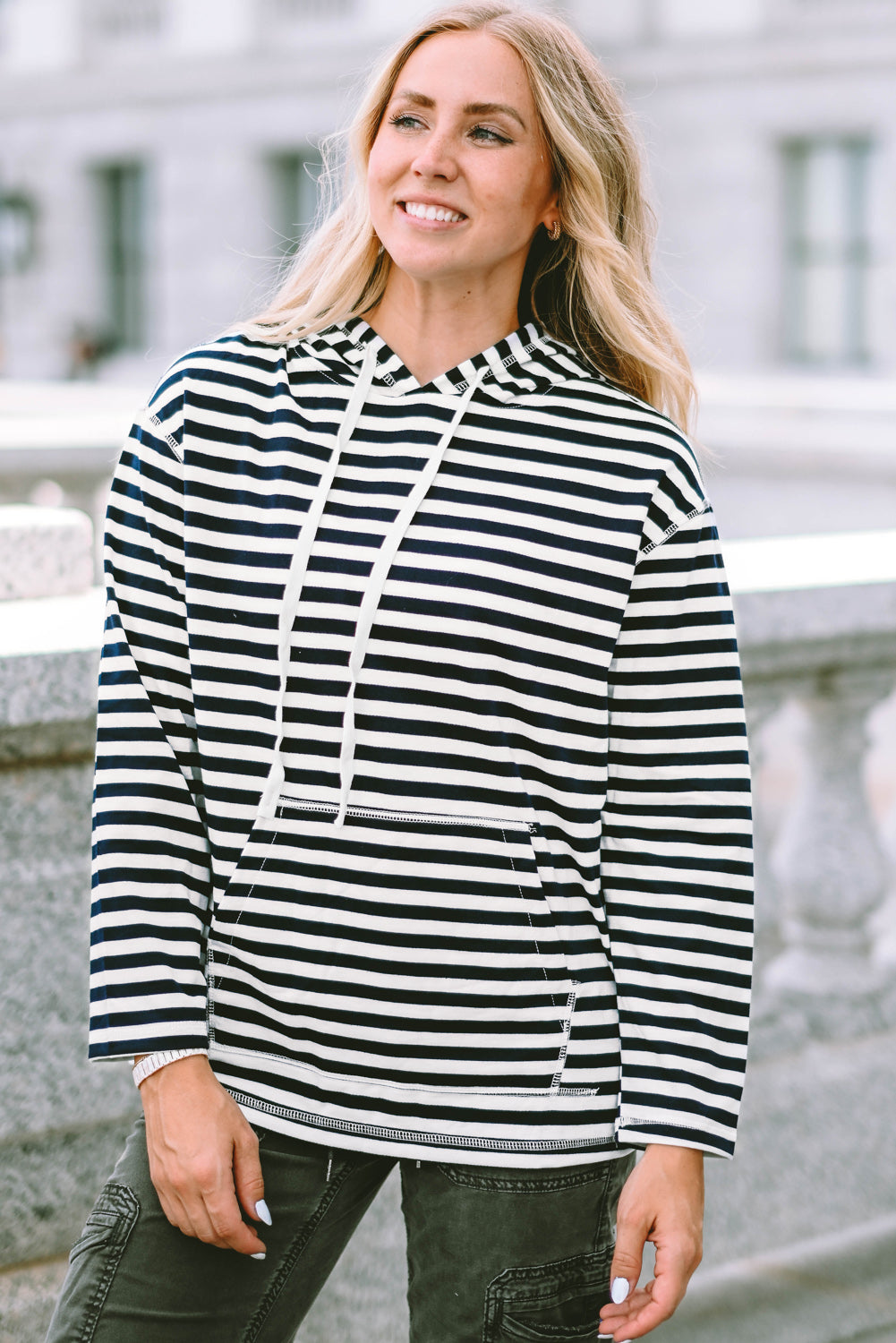 A person with long, blonde hair wearing a trendy Stripe Kangaroo Pocket Drawstring Hoodie with black and white stripes and dark green pants is standing outdoors, facing away from the camera.