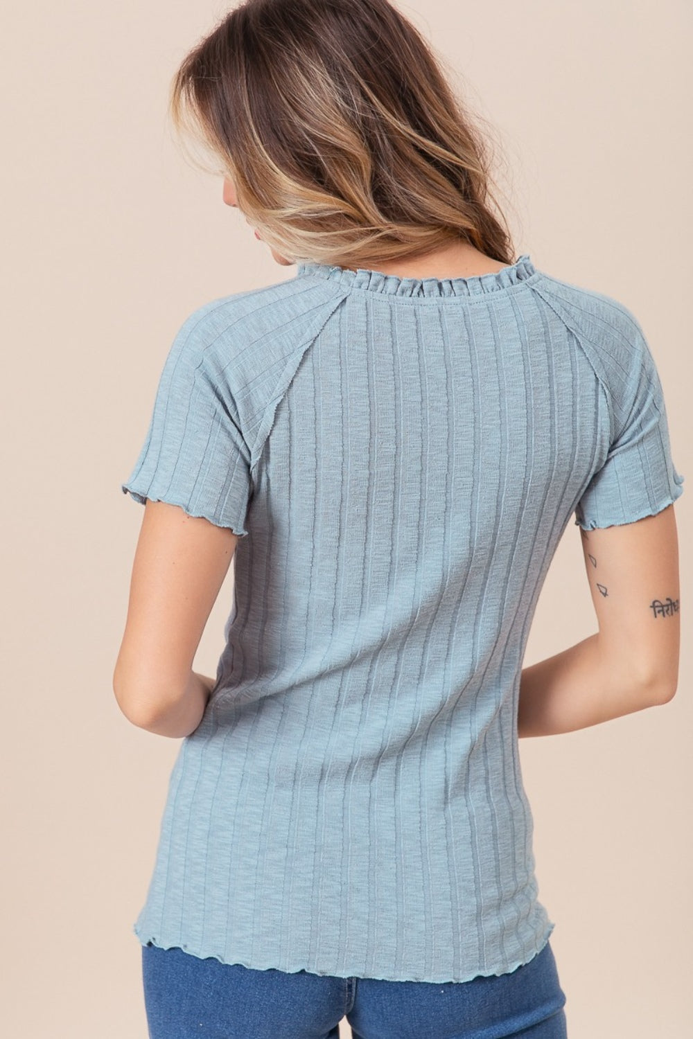 A person wearing the BiBi Ruffle Round Neck Merrow Edge Rib Top, featuring short sleeves and a light blue hue, stands against a neutral background. They have long, wavy hair, with tattoos adorning their arm.