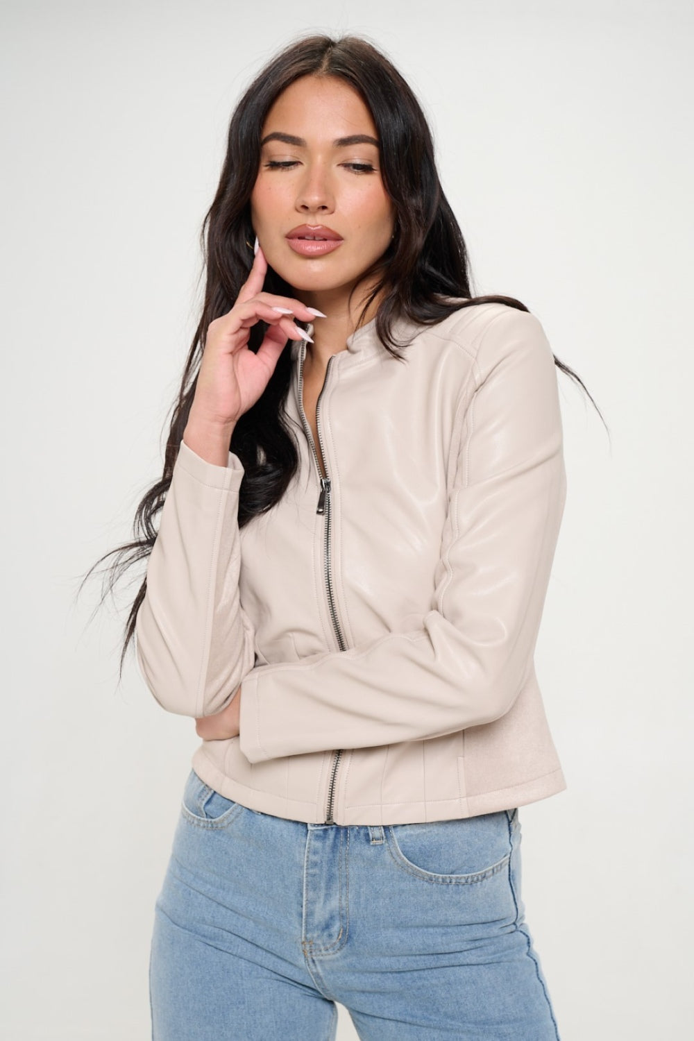 A woman with long dark hair, wearing the cruelty-free Coalition LA Zip Up Vegan Moto Jacket in light beige, pairs it with blue jeans as she stands with her arms crossed.