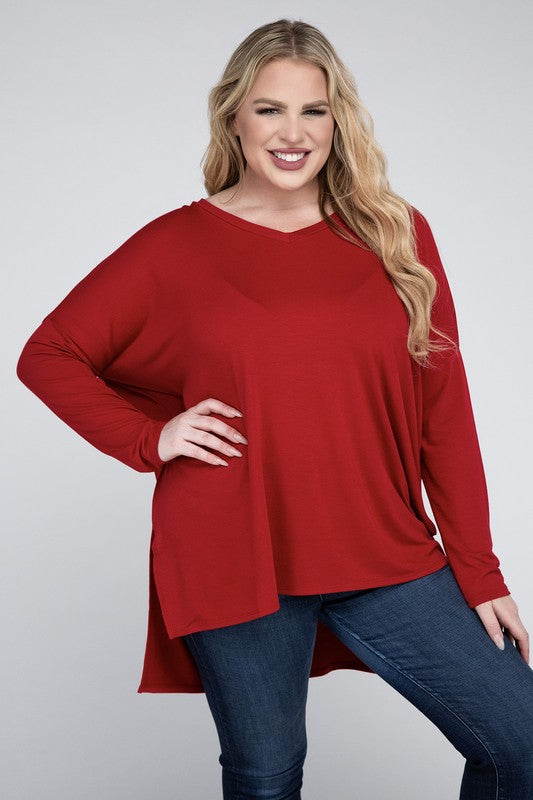 A woman with long, blonde hair wears a Plus Dolman Sleeve V-Neck Side Slit Hi-Low Hem Top and blue jeans, posing with one hand on her hip and smiling at the camera.